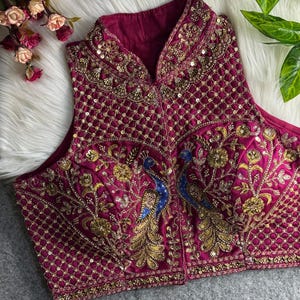 May include: A sleeveless, magenta-colored top with intricate gold embroidery and sequin detailing. The design features two peacocks with blue and gold accents. The top has a high collar and is likely part of a formal wear collection.