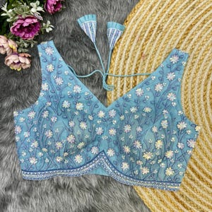 May include: A light blue embroidered blouse with a V-neckline and floral pattern. The blouse has a tie closure at the back and is decorated with sequins.