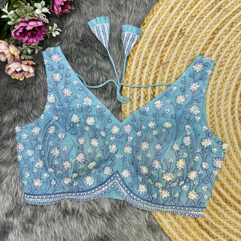 May include: A light blue embroidered blouse with a V-neckline and floral pattern. The blouse has a tie closure at the back and is decorated with sequins.
