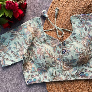 Pode incluir: Uma blusa floral azul clara com mangas curtas. A blusa apresenta um decote em V e um delicado padrão floral em tons de azul, roxo e dourado. A blusa é amarrada nas costas com um cordão a condizer.