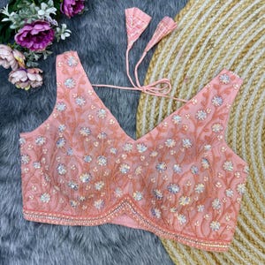 May include: A pink embroidered blouse with sequins and a V-neckline. The blouse has a tie closure at the back and is decorated with floral patterns.