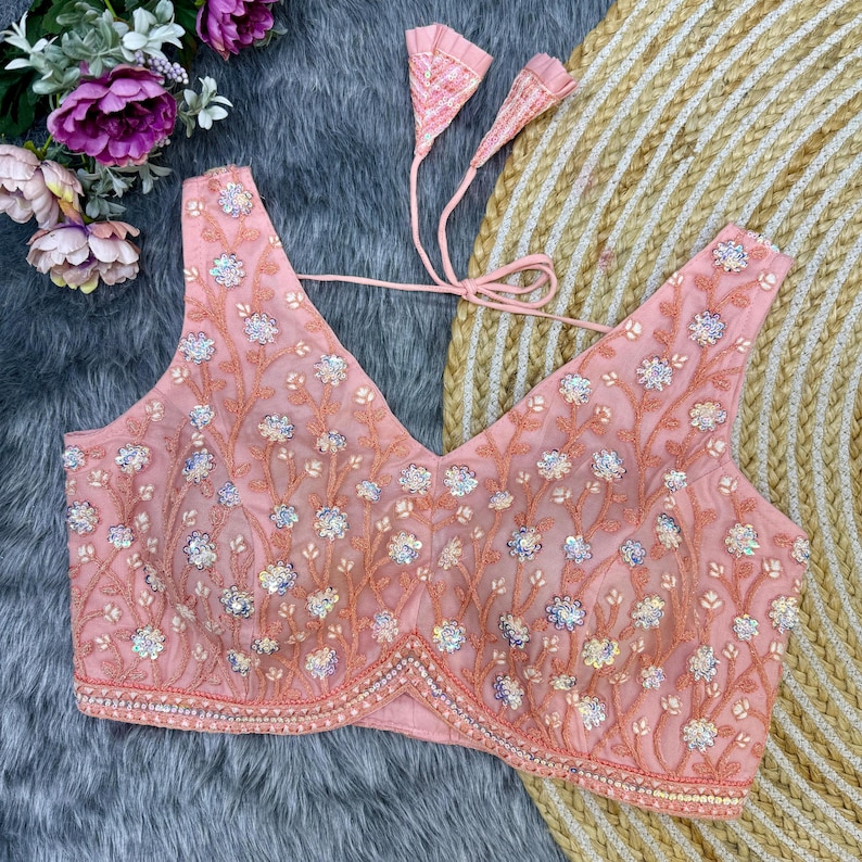May include: A pink embroidered blouse with sequins and a V-neckline. The blouse has a tie closure at the back and is decorated with floral patterns.