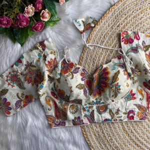 Pode incluir: Blusa cor marfim com um vibrante padrão floral paisley em tons de vermelho, laranja, roxo e verde-azulado. A blusa apresenta mangas curtas e decote em coração. Um acessório a condizer também está incluído.