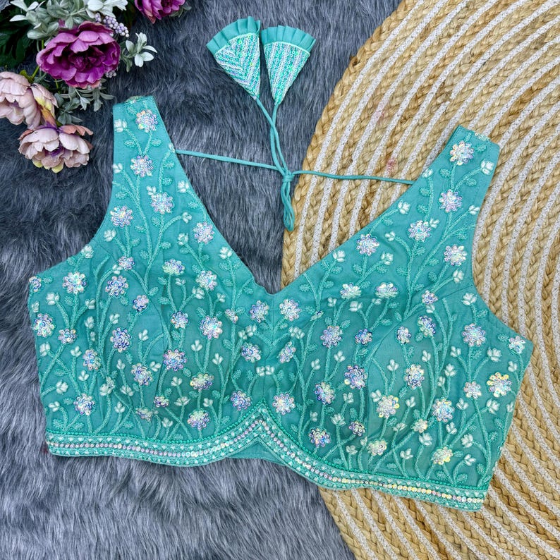 May include: A teal blue embroidered blouse with a V-neckline and a floral pattern. The blouse has a beaded trim and a tie closure at the back.