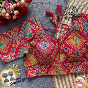Pode incluir: Uma blusa rosa vibrante bordada com intrincados fios dourados e padrões florais coloridos. A blusa apresenta um decote em V e mangas curtas. O texto "The Traditional Street" é visível. Há também um distintivo de "Premium Quality".