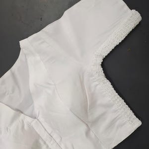 May include: A solid white blouse featuring short sleeves with a textured, gathered, and ruffled trim along the sleeve edges. The garment has a V-neckline and structured seams running vertically down the front, creating a clean and simple appearance.