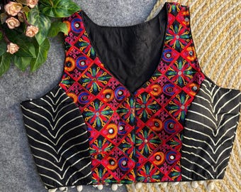 May include: A sleeveless, black vest with a V-neck and colorful embroidery. The vest features a red base with geometric patterns in blue, green, and yellow, accented with small mirrors. Black and white striped panels are on the sides, and white tassels line the bottom.