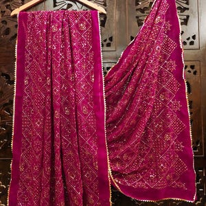 May include: A magenta-colored scarf with an intricate gold pattern and a scalloped gold trim. The scarf is draped over a wooden hanger, showcasing its detailed design and texture. The background is a dark wooden door.