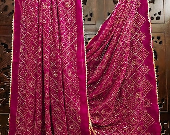 Può includere: Una sciarpa magenta con un intricato motivo dorato e un bordo dorato smerlato. La sciarpa è drappeggiata su una gruccia di legno, mostrando il suo design dettagliato e la sua consistenza. Lo sfondo è una porta di legno scuro.