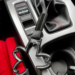 Könnte beinhalten: Ein schwarzer Innenraum eines Autos mit einem schwarzen Lederschalthebelbalg und einem schwarzen Schalthebelknauf. Ein schwarzes Autozubehör mit einem Schmetterlingsdesign ist am Schalthebelknauf befestigt.