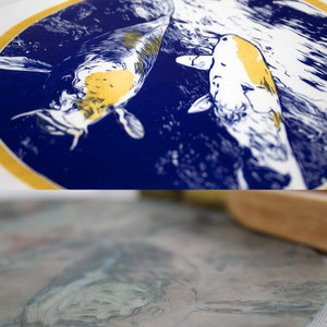May include: A blue and gold print of two koi fish swimming in a circle. The fish are white with gold markings and are swimming in a blue circle with a gold border.