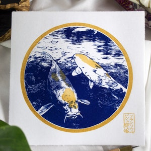 May include: A blue and gold print of two koi fish swimming in a circle. The print is on white paper and has a gold seal with Japanese characters in the bottom right corner.