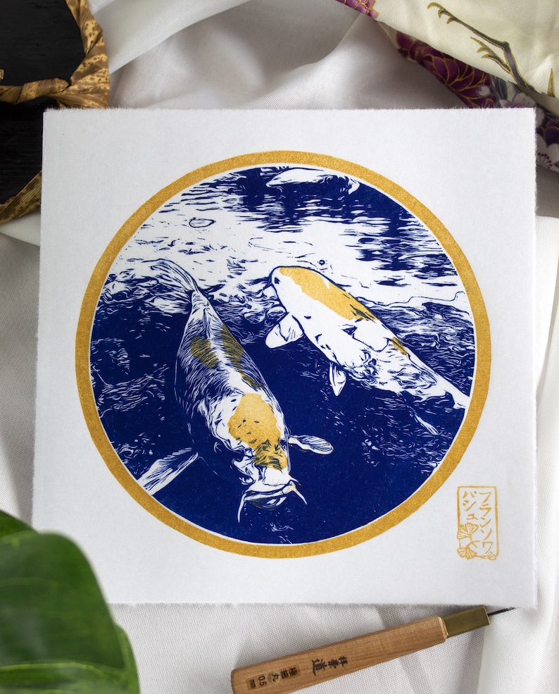 May include: A blue and gold print of two koi fish swimming in a circle. The print is on white paper and has a gold seal with Japanese characters in the bottom right corner.