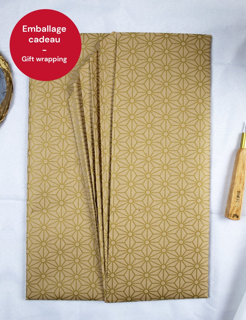 May include: A stack of brown gift wrapping paper with a gold geometric pattern. The paper is folded in a fan shape and has the text "Emballage cadeau - Gift wrapping" printed on the top sheet.