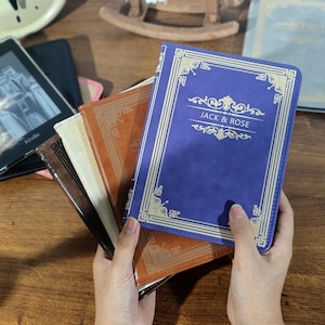 May include: A collection of book-style Kindle covers in various colors, including blue, brown, and tan. The blue cover features the text "JACK & ROSE" in gold lettering. Other covers are visible, suggesting a range of options for e-readers.