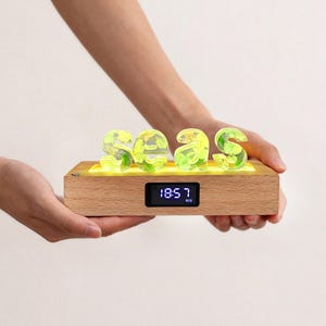 Può includere: Un blocco di legno con la scritta "2023" in lettere verdi traslucide. Il blocco ha un orologio digitale che mostra "18:57". Le lettere sono illuminate da una luce gialla, creando un pezzo decorativo moderno.