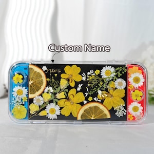 May include: A clear Switch console case with a black center panel featuring dried yellow flowers and orange slices. The case has blue and red sides, with additional floral accents. The word "Flora" is printed on the case.