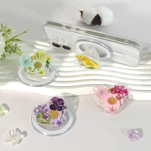 Puede incluir: Soportes para teléfono en forma de corazón con flores secas en resina transparente. Uno de los soportes tiene la palabra "FLY". Los soportes están sobre una superficie blanca con una funda transparente para teléfono adjunta a uno. Las flores son amarillas, rosas, moradas y azules.