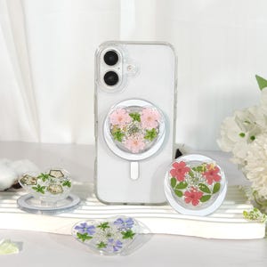 May include: Clear phone case with a heart-shaped pop socket featuring pink and white dried flowers. Additional heart-shaped pop sockets with dried flowers in various colors are also displayed.
