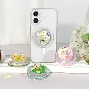 Puede incluir: Una funda transparente para teléfono con un diseño floral y un soporte para teléfono a juego. El soporte para teléfono tiene la palabra "Fly" y un diseño floral. Hay otros tres soportes para teléfono con diseños florales y las iniciales "DW", "R+D" y "ws".