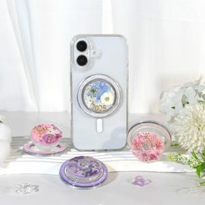 May include: Clear phone case with a floral design and a magnetic ring holder. The ring holder features a mix of dried flowers and the number "2025". Additional floral ring holders in pink and purple are also displayed.