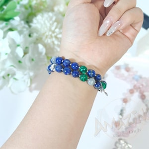 Pode incluir: Duas pulseiras de contas com pedras preciosas azuis, verdes e brancas são usadas no pulso. As pulseiras são feitas de contas redondas. O fundo inclui flores brancas e o texto "Amazing Flowers Crafts".