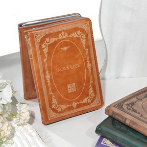 Puede incluir: Un libro de cuero marrón con el texto "JACK & ROSE" en el centro. La portada presenta un diseño dorado ornamentado con una corona en la parte superior. Otros libros de varios colores son visibles en el fondo.
