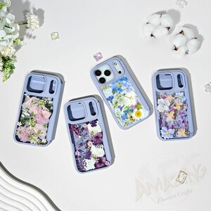 Puede incluir: Cuatro fundas de teléfono lavanda con diseños florales. Cada funda presenta una disposición diferente de flores secas en tonos rosa, morado, azul y blanco. Las fundas están dispuestas sobre una superficie blanca con algodón y elementos decorativos.