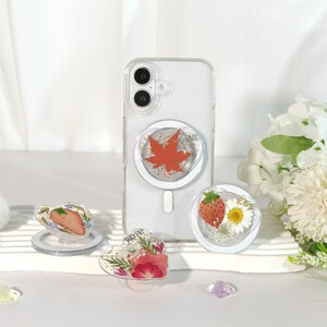 Pode incluir: Uma capa de telefone transparente com um suporte em forma de coração com uma folha de bordo vermelha. Suportes adicionais em forma de coração exibem um morango, margaridas e rosas. As pegas são transparentes com desenhos florais e de frutas.
