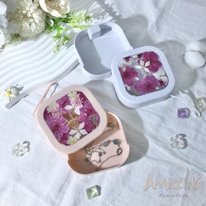 Puede incluir: Dos cajas de joyería cuadradas con diseños florales. La parte superior de las cajas es transparente y muestra flores prensadas. La parte inferior de las cajas es rosa y blanca. Las cajas están abiertas y muestran un collar en el interior.