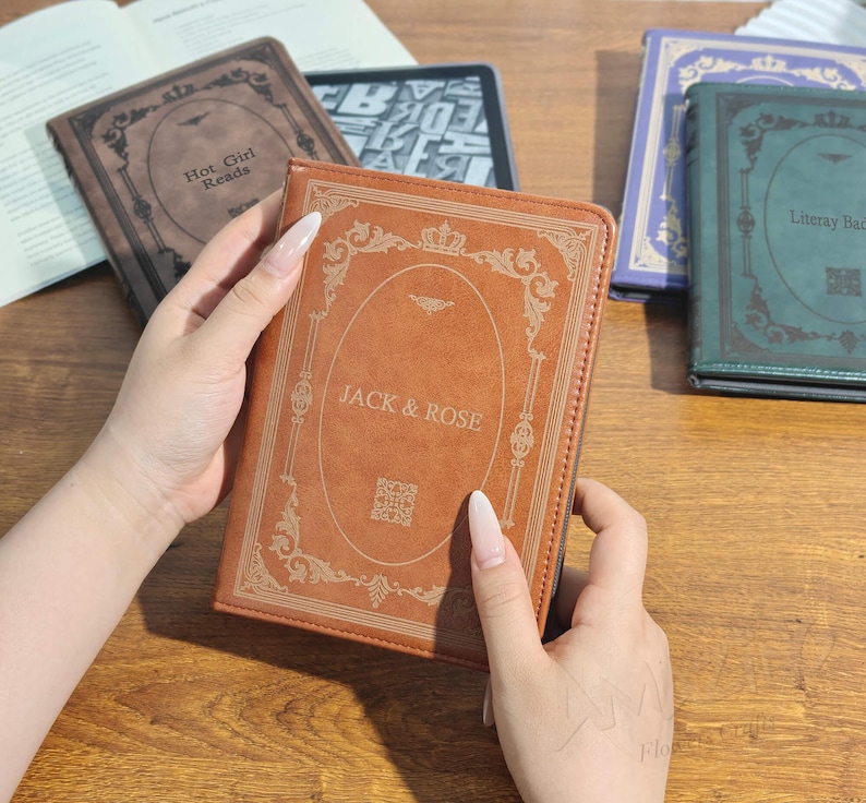 Puede incluir: Una funda de libro de cuero marr&oacute;n con el texto "JACK & ROSE" en letras doradas. La funda tiene un dise&ntilde;o ornamental en los bordes y un peque&ntilde;o c&oacute;digo QR. Se ven otras fundas de libros, incluida una con el texto "Hot Girl Reads".