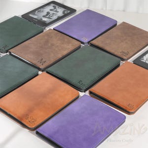 May include: A selection of personalised e-reader covers in shades of green, brown, orange, and purple. Each cover displays a name or initials, with a Kindle e-reader visible in the background. The covers are arranged on a white surface.