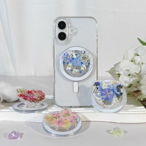 May include: A clear phone case with a heart-shaped phone grip containing dried flowers and the name "Xie". Other heart-shaped grips with floral designs are shown, alongside a clear phone case.