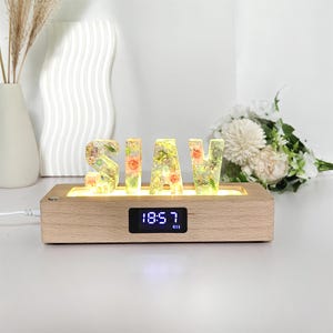 Puede incluir: Base de madera con las letras S, H, A e Y, cada una llena de flores y vegetación coloridas. La base tiene un reloj digital que muestra las 18:57. Las letras están iluminadas con un brillo cálido.