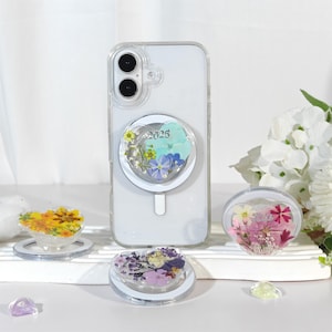 May include: Clear phone case with a heart-shaped pop socket featuring dried flowers and the year "2025". Additional heart-shaped pop sockets with floral designs are displayed. The phone case is on a white surface.