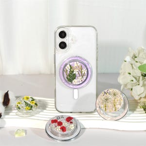 Pode incluir: Uma capa de telefone transparente com um suporte floral roxo que mostra flores secas. São exibidos três suportes florais adicionais com diferentes arranjos florais. Os suportes de telefone têm um design claro e circular.