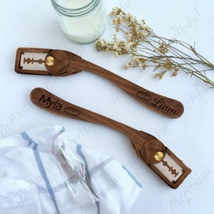 May include: Two wooden bread lame tools with engraved names "Myla" and "Liam". Each tool has a blade holder with a gold-colored fastener. The handles are dark brown with a bread loaf design. A jar of milk, flowers, and a white and blue striped cloth are in the background.