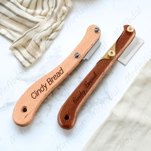 Könnte beinhalten: Zwei Brotschneidewerkzeuge aus Holz, eines hell und eines dunkel, jeweils mit einer Klinge und dem eingravierten Text "Cindy Bread". Das helle Holzwerkzeug hat silberne Schrauben, während das dunkle Holzwerkzeug Messingakzente hat.