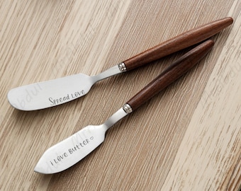 May include: Two butter knives with stainless steel blades and wooden handles. One knife has the text "Spread Love" and the other says "I Love Butter". The knives are on a wooden surface.