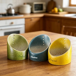 May include: Three ceramic utensil holders in green, teal, and yellow, each with a name: Rachel, Martina, and Fraisel. The holders have a textured finish and a curved design, with small holes near the top. They sit on a wooden surface.