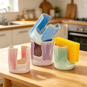 May include: Several ceramic sponge holders in shades of pink, purple, blue, and green. Each has a textured surface and a slot for a sponge. The names "Rachel", "Sulis", "Raisel", and "Martina" are printed on the holders, which are displayed on a wooden surface.