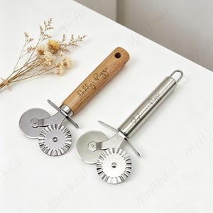 May include: Two pizza cutters with dual blades, one smooth and one fluted. One cutter has a light wood handle engraved with "Daddy's Pizza" and the other is entirely stainless steel with "Stephanie's Pizza" engraved on the handle.