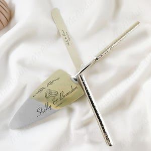 Peut inclure: Un ensemble de couteau et de pelle à gâteau argentés. La pelle porte les noms "Shelly & Brandon" et la date "10.16.24". Le couteau a un manche texturé et une gravure assortie.