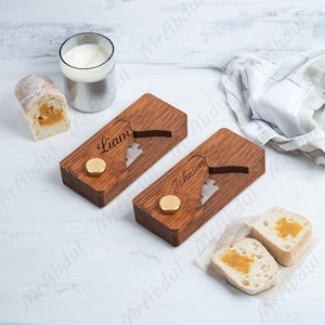 Könnte beinhalten: Zwei Brotschneider aus Holz mit den eingravierten Namen "Liam" und "Johnson". Jeder Schneider hat einen goldfarbenen Knopf und einen brotförmigen Ausschnitt. Geschnittenes Brot mit Marmelade und ein Glas Milch sind ebenfalls vorhanden.