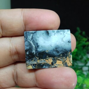 Puede incluir: Una piedra preciosa cuadrada con un patr&oacute;n de paisaje. La piedra presenta una mezcla de colores, incluyendo blanco, negro, gris y marr&oacute;n. El patr&oacute;n se asemeja a un cielo nublado sobre un paisaje oscuro. La piedra se sostiene en una mano.