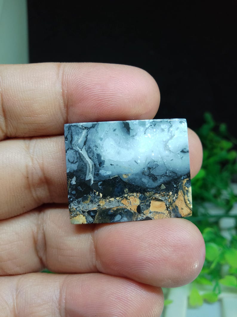 Puede incluir: Una piedra preciosa cuadrada con un patr&oacute;n de paisaje. La piedra presenta una mezcla de colores, incluyendo blanco, negro, gris y marr&oacute;n. El patr&oacute;n se asemeja a un cielo nublado sobre un paisaje oscuro. La piedra se sostiene en una mano.