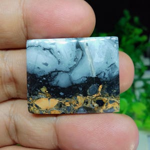 Puede incluir: Una piedra preciosa cuadrada con un patr&oacute;n &uacute;nico. La parte superior es azul claro con vetas grises oscuras, que pasa a negro y luego a una mezcla de oro y marr&oacute;n en la parte inferior. La piedra se sostiene en una mano.