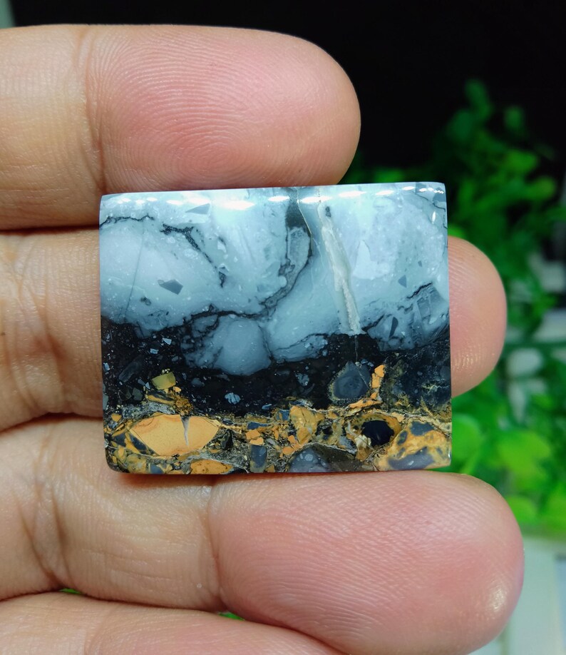 Puede incluir: Una piedra preciosa cuadrada con un patr&oacute;n &uacute;nico. La parte superior es azul claro con vetas grises oscuras, que pasa a negro y luego a una mezcla de oro y marr&oacute;n en la parte inferior. La piedra se sostiene en una mano.
