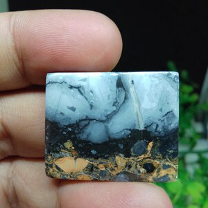 Puede incluir: Una piedra preciosa cuadrada con una apariencia en capas. La capa superior es azul claro con vetas blancas, seguida de una banda negra y una capa inferior marr&oacute;n y dorada. La piedra se sostiene entre los dedos.