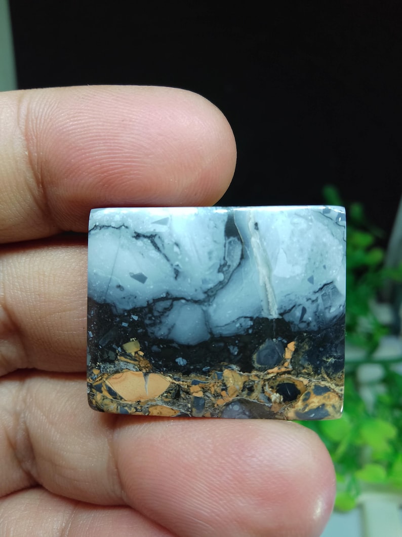 Puede incluir: Una piedra preciosa cuadrada con una apariencia en capas. La capa superior es azul claro con vetas blancas, seguida de una banda negra y una capa inferior marr&oacute;n y dorada. La piedra se sostiene entre los dedos.